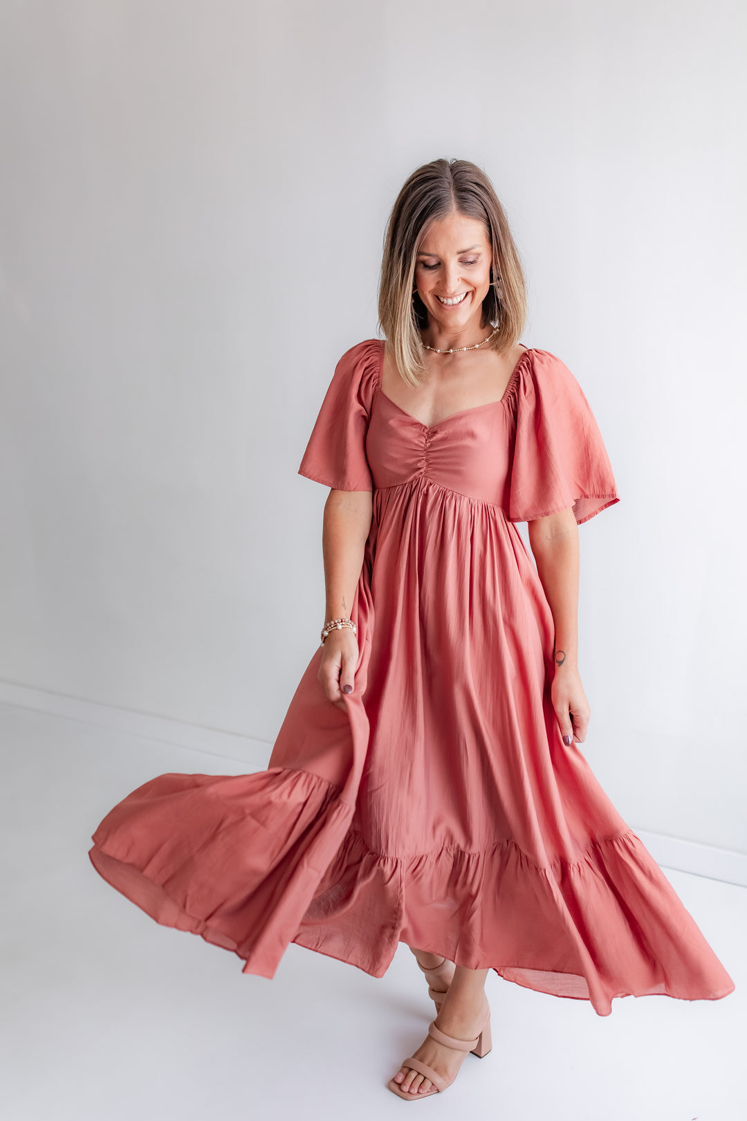 A woman wears a cinnamon-colored dress with a smocked back, a sweetheart bodice, and flutter sleeves, styled with boots.
