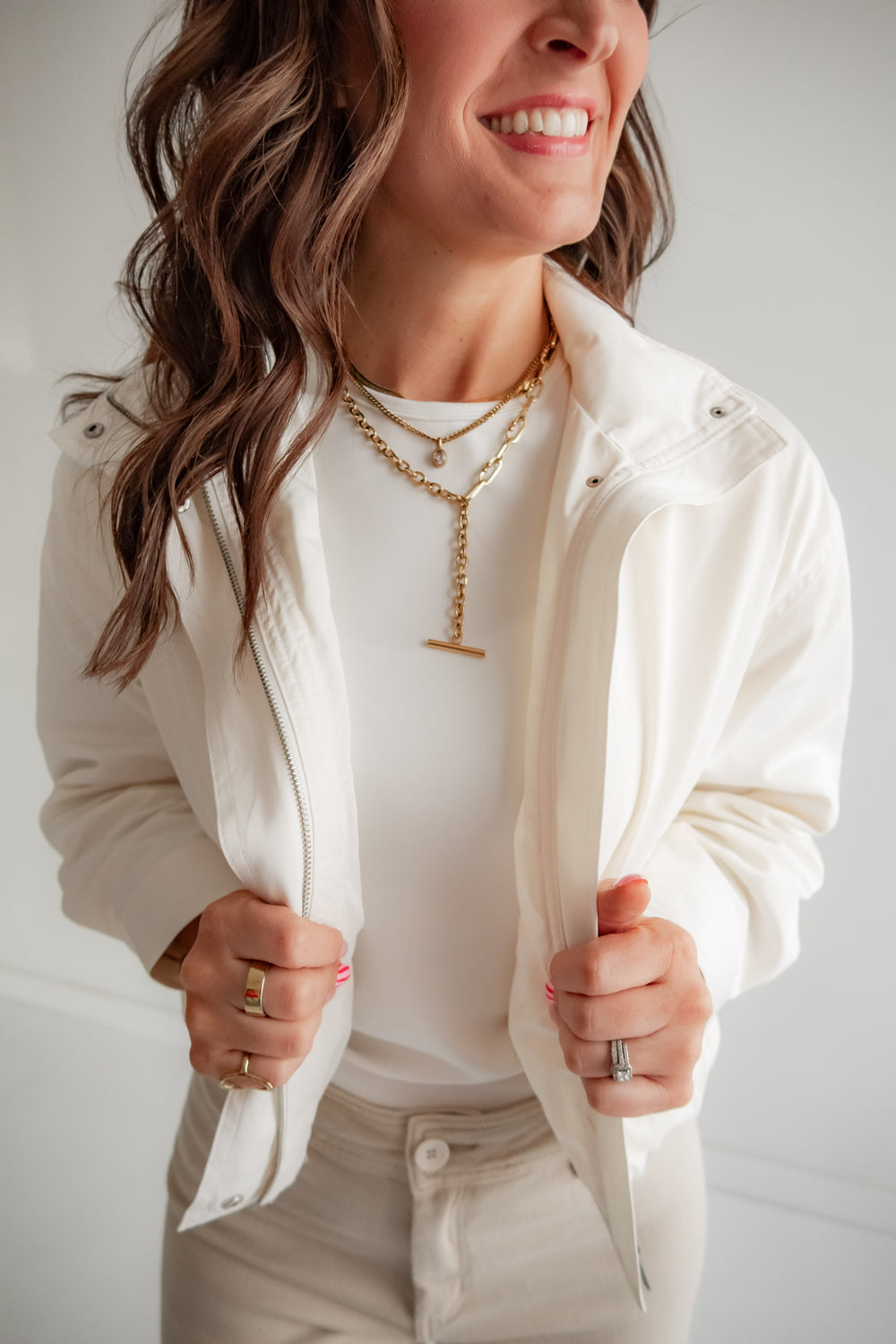 Woman wearing a white jacket over a white shirt with layered necklaces against a neutral background