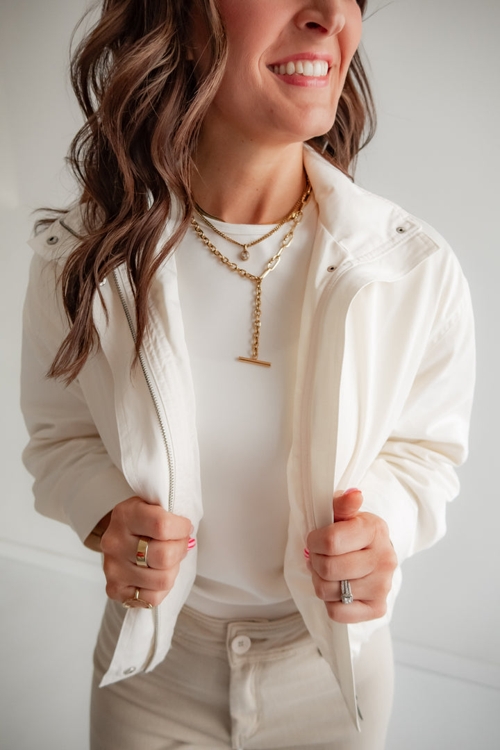 Woman wearing a white jacket over a white shirt with layered necklaces against a neutral background