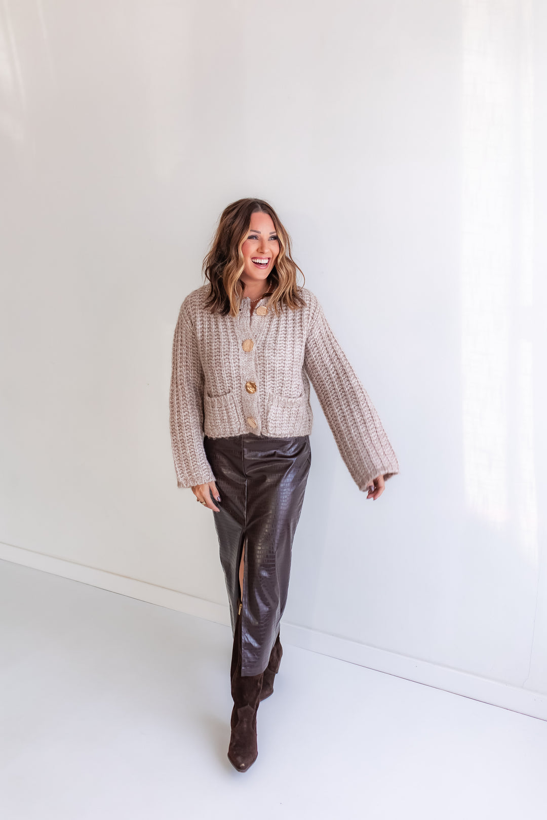 Woman wearing a taupe cardigan and brown leather midi skirt against a white background