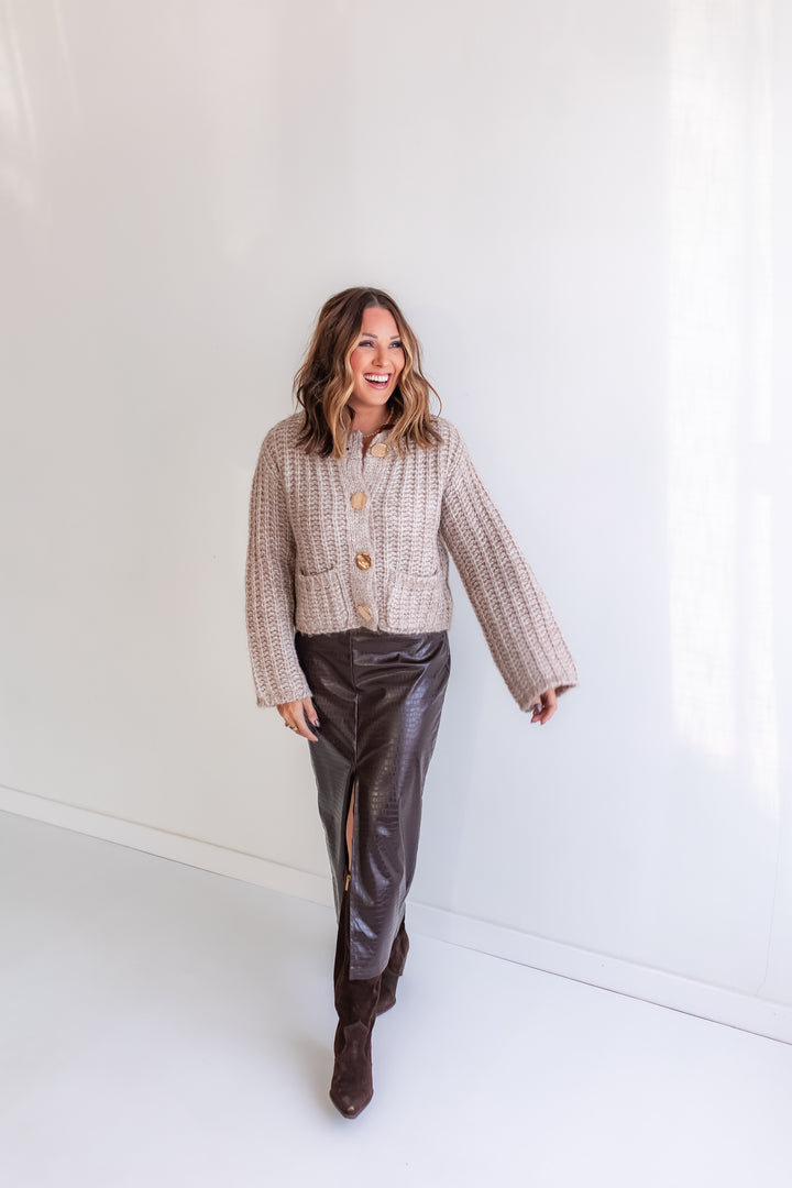 Woman wearing a taupe cardigan and brown leather midi skirt against a white background