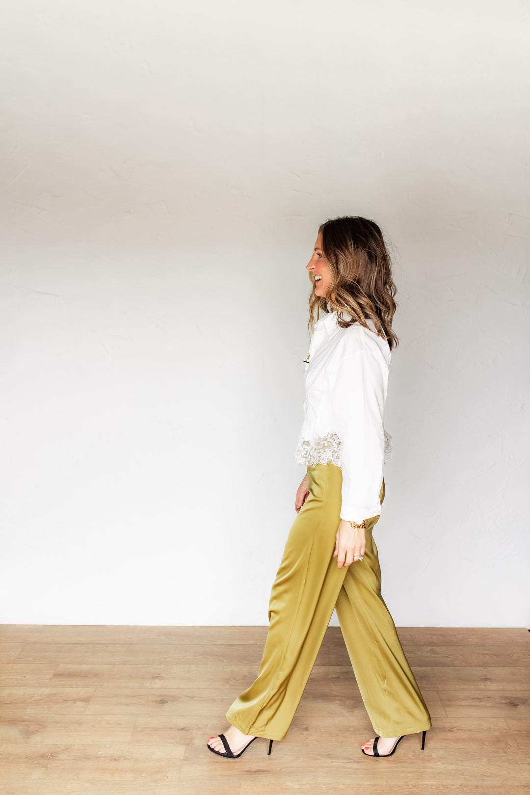 Sideview of woman walking wearing olive gold satin wide leg pants with a white lace-trim blouse